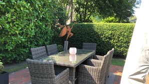 A wicker outdoor dining set with a glass table and chairs, surrounded by greenery and a brick patio. - Gemütliche 2,5 Zimmer unter Reet in SPO (Sankt Peter-Ording)