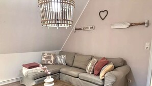 A cozy living room with a sectional sofa, a wooden coffee table, a hanging light fixture, and a wall-mounted sign reading "BEACH BAR". - Gemütliche 2,5 Zimmer unter Reet in SPO (Sankt Peter-Ording)