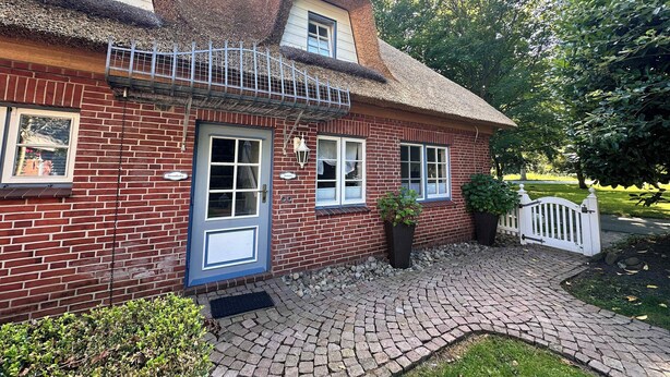 A brick house with a thatched roof, a white picket fence, and a paved driveway. - Gemütliche 2,5 Zimmer unter Reet in SPO (Sankt Peter-Ording)