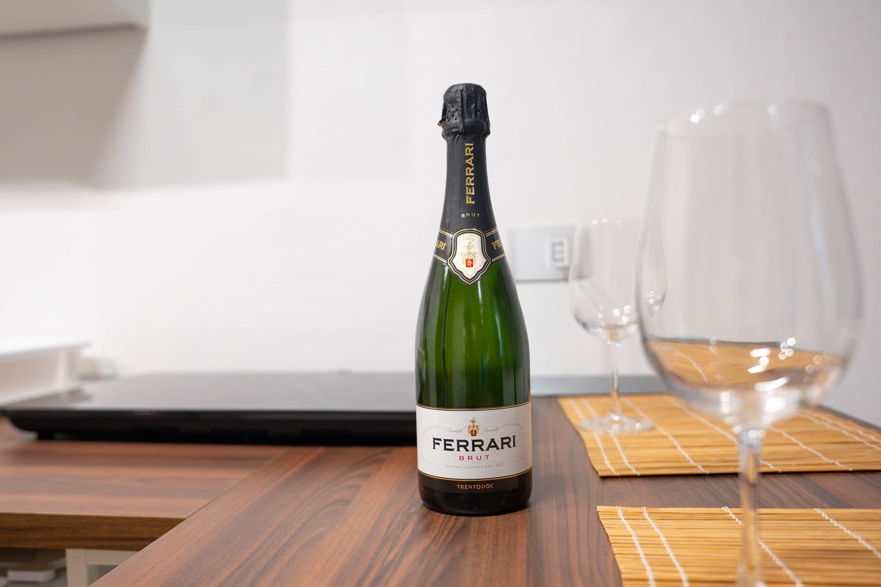 A bottle of Ferrari Brut wine on a wooden table with a glass of wine and a laptop in the background.