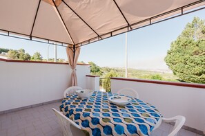 Un balcón cubierto con una mesa y sillas, vista al campo y un árbol grande.