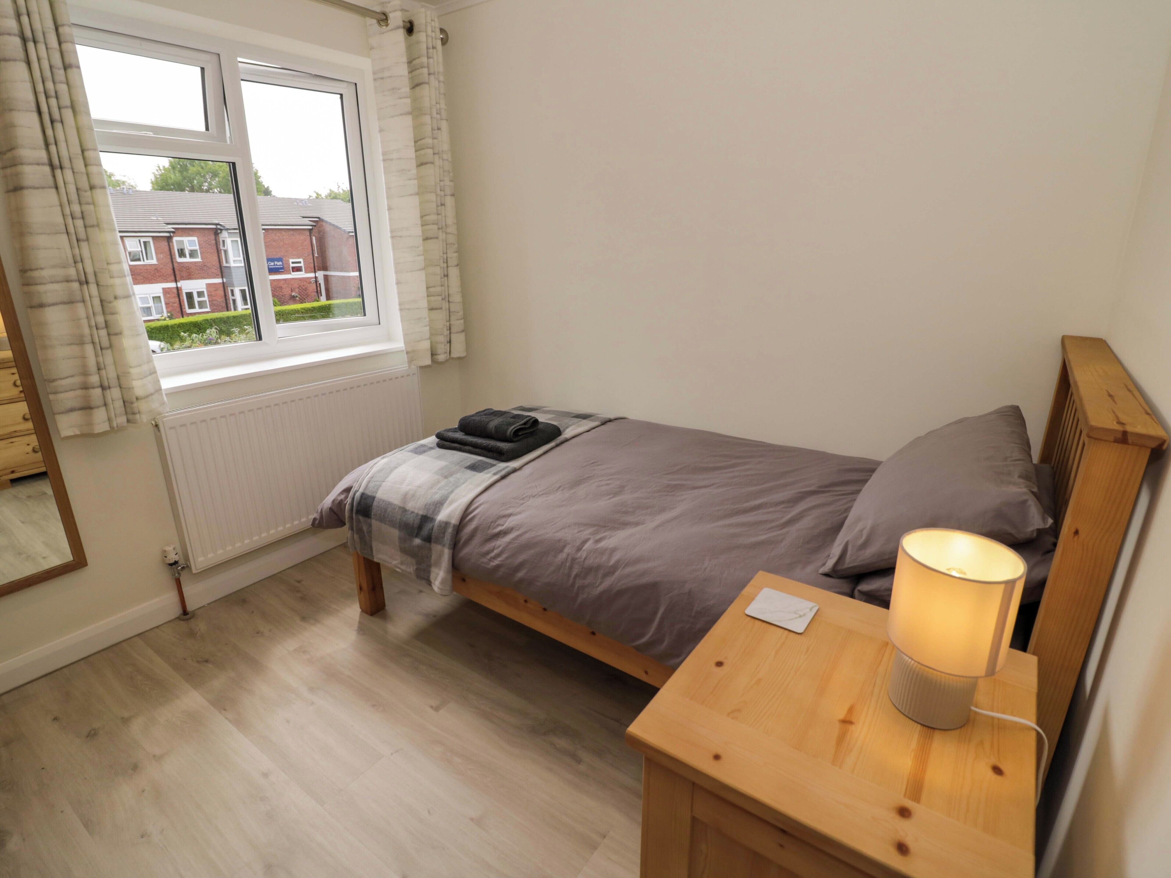Une petite chambre à coucher, bien rangée, avec un lit simple, une table de chevet avec une lampe, une fenêtre avec des rideaux et une vue sur un bâtiment en briques à l’extérieur.