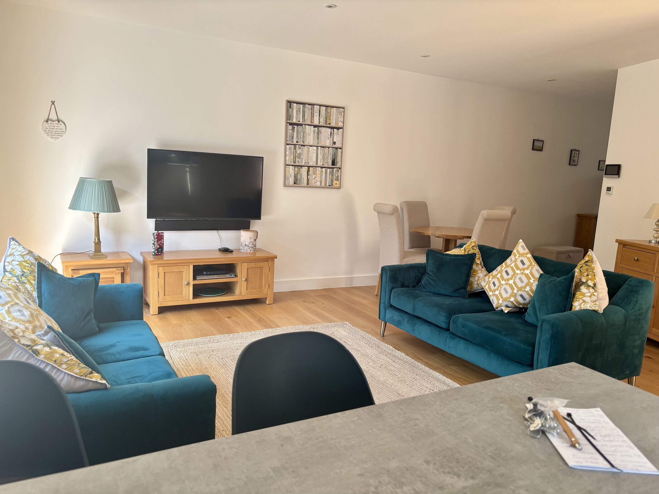 A modern living room with a flat-screen TV, a wooden entertainment unit, teal sofas, a dining table, and a small wooden cabinet.