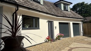 A house with a sloped roof, two windows, a door, and a garage. There are potted plants and gravel in the driveway. - The Retreat (Ferndown)