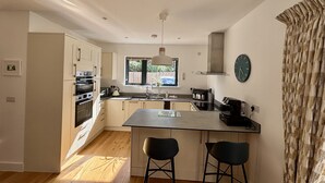 A modern kitchen with white cabinets, a central island with two stools, a window with a view, and a clock on the wall. - The Retreat (Ferndown)