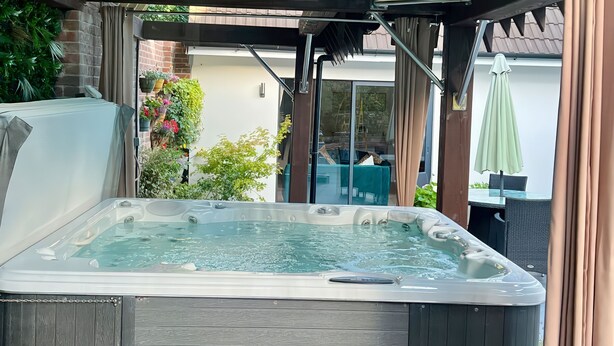 A hot tub with a wooden exterior, surrounded by a covered patio area with a glass door leading to an outdoor seating area. - The Retreat (Ferndown)