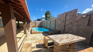 An outdoor patio area with a pool, wooden furniture, and a stone wall. - Casa em Natal com Piscina a 290m da Praia (Natal)