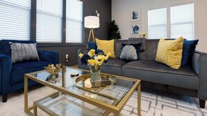 A modern living room with a blue sofa, a gray sectional, and a glass coffee table with a gold frame. - 4BR Retreat for 12 w Balcony, Mins to Broadway! (Nashville)