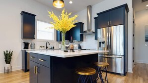 A modern kitchen with dark cabinetry, a large island with stools, stainless steel appliances, and a vase of yellow flowers. - 4BR Retreat for 12 w Balcony, Mins to Broadway! (Nashville)