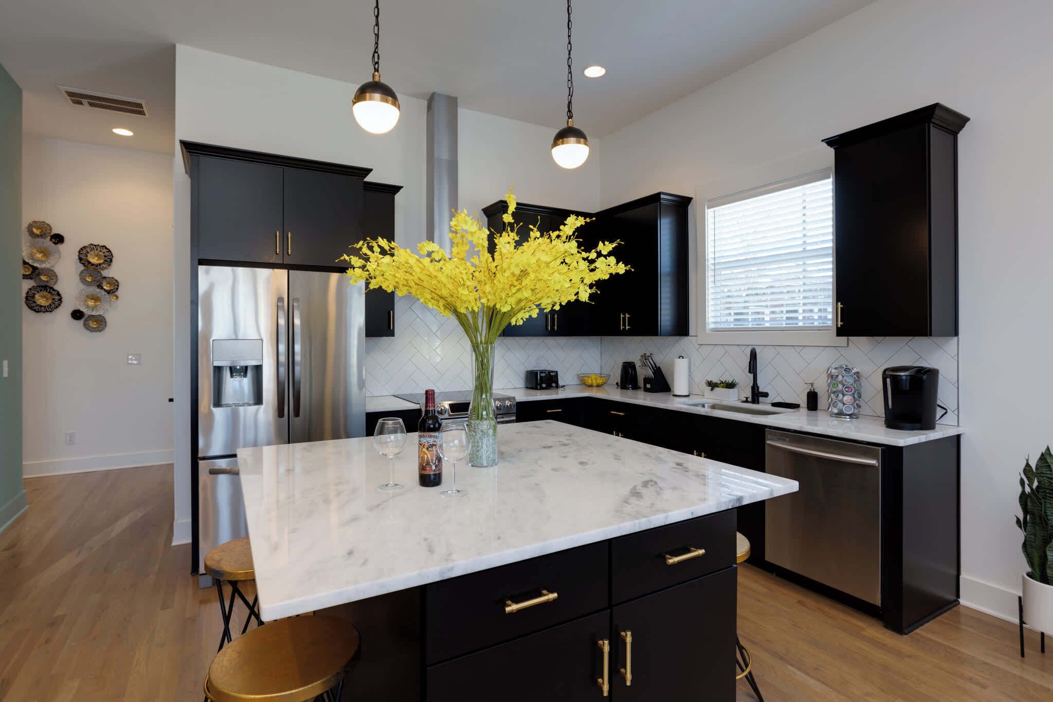 Une cuisine moderne avec des meubles noirs, un îlot en marbre, des appareils électroménagers en acier inoxydable et un vase rempli de fleurs jaunes.