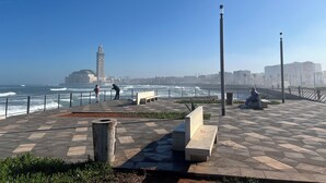 Un paseo marítimo con bancos, un contenedor de basura y una vista despejada del mar y edificios lejanos.