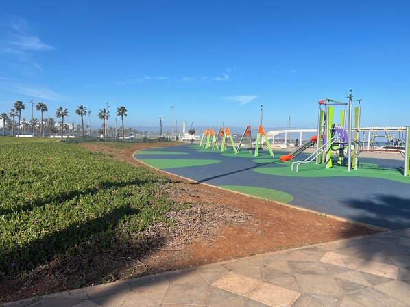 Un parque infantil con diversas estructuras de juego, un sendero pavimentado y una zona verde con palmeras al fondo.