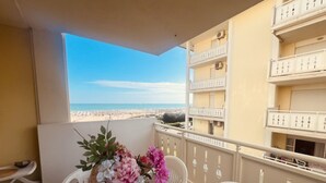 Un balcón con una mesa, sillas y un florero. El balcón tiene vista a una playa y edificios cercanos.