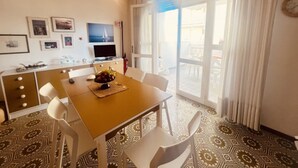 Un comedor con una mesa de madera, sillas blancas, un frutero y un televisor sobre un mueble.