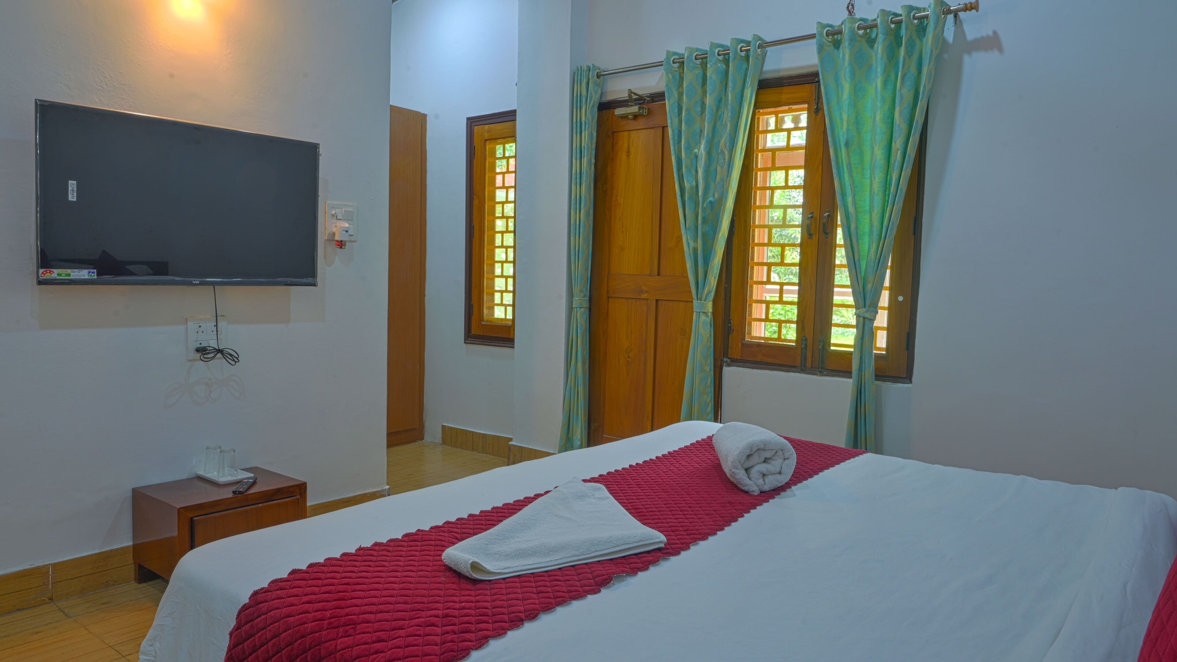 Una habitación de hotel con una cama, un televisor montado en la pared, una mesita de noche con un teléfono y una ventana con cortinas.