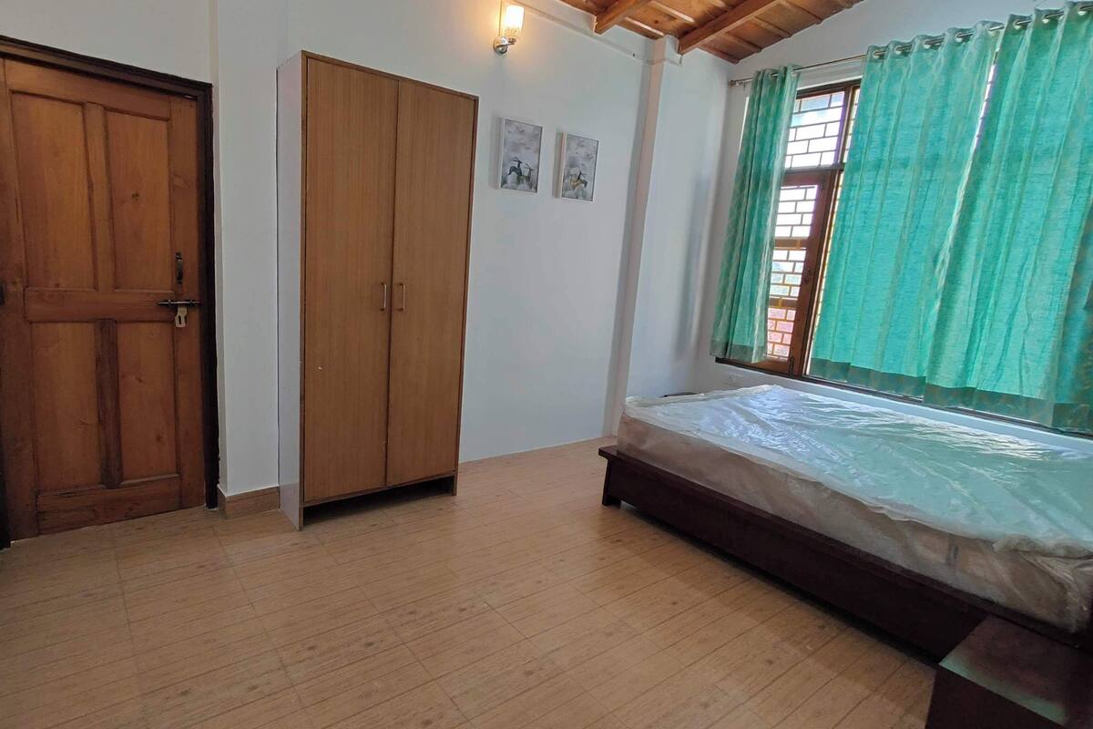 A room with a wooden door, a wardrobe, a bed with a mattress, a wall-mounted TV, and a window with curtains.