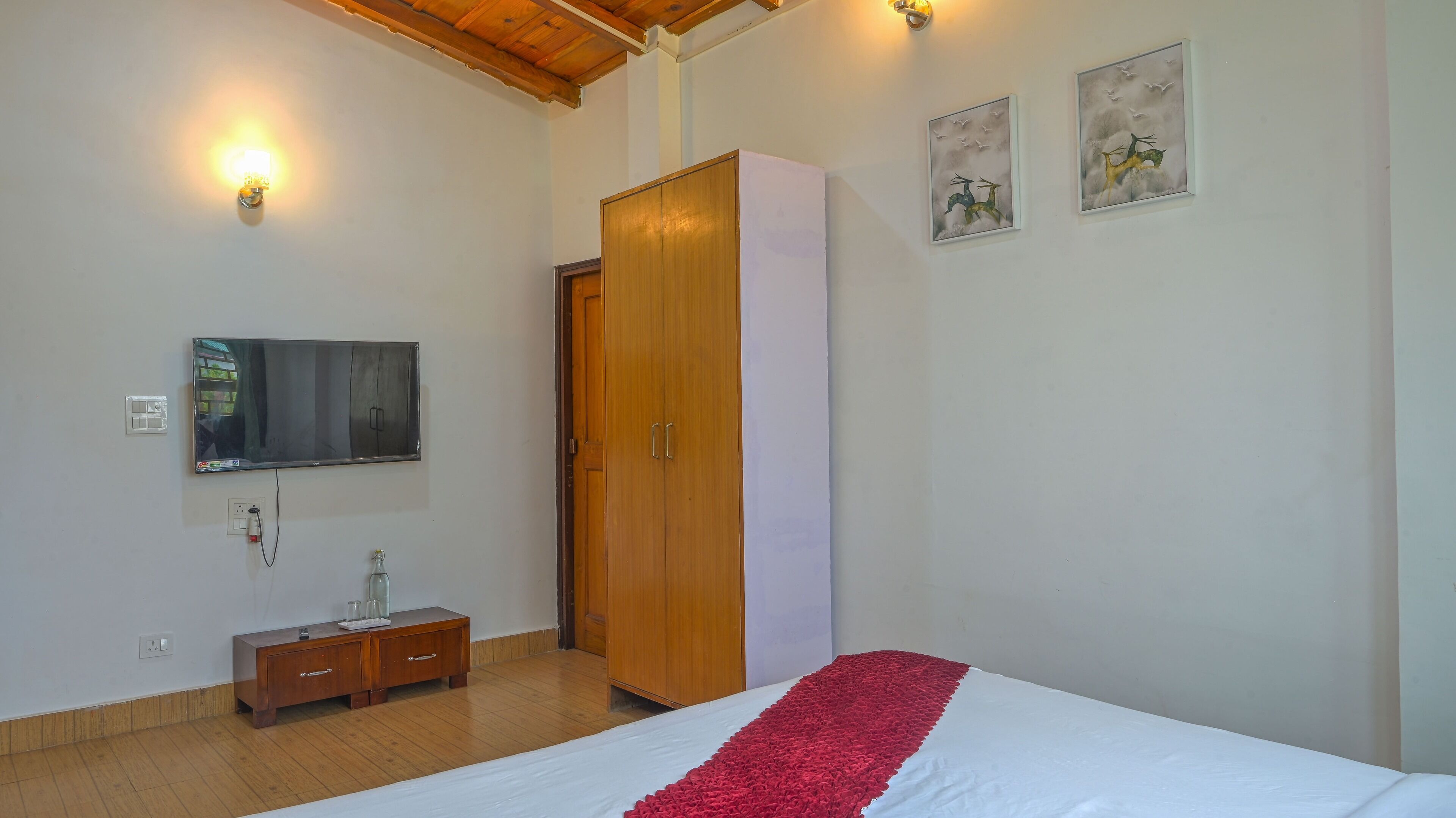 Une chambre à coucher avec un lit, une armoire en bois, un téléviseur fixé au mur et une petite table de chevet avec un verre.