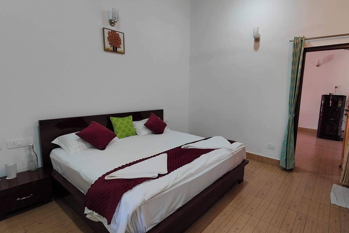 A neatly made bed with pillows, a bedside table, a wall-mounted light, and a door with a curtain.