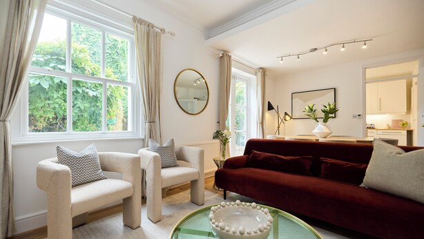 Ein Wohnzimmer mit einem roten Sofa, zwei Sesseln, einem Glastisch und einem großen Fenster mit Vorhängen.
