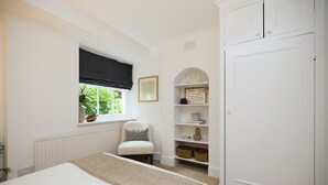 Ein Schlafzimmer mit Bett, Fenster mit Karos, einem Sessel, einer Kommode mit Dekoration und einem weißen Kleiderschrank.