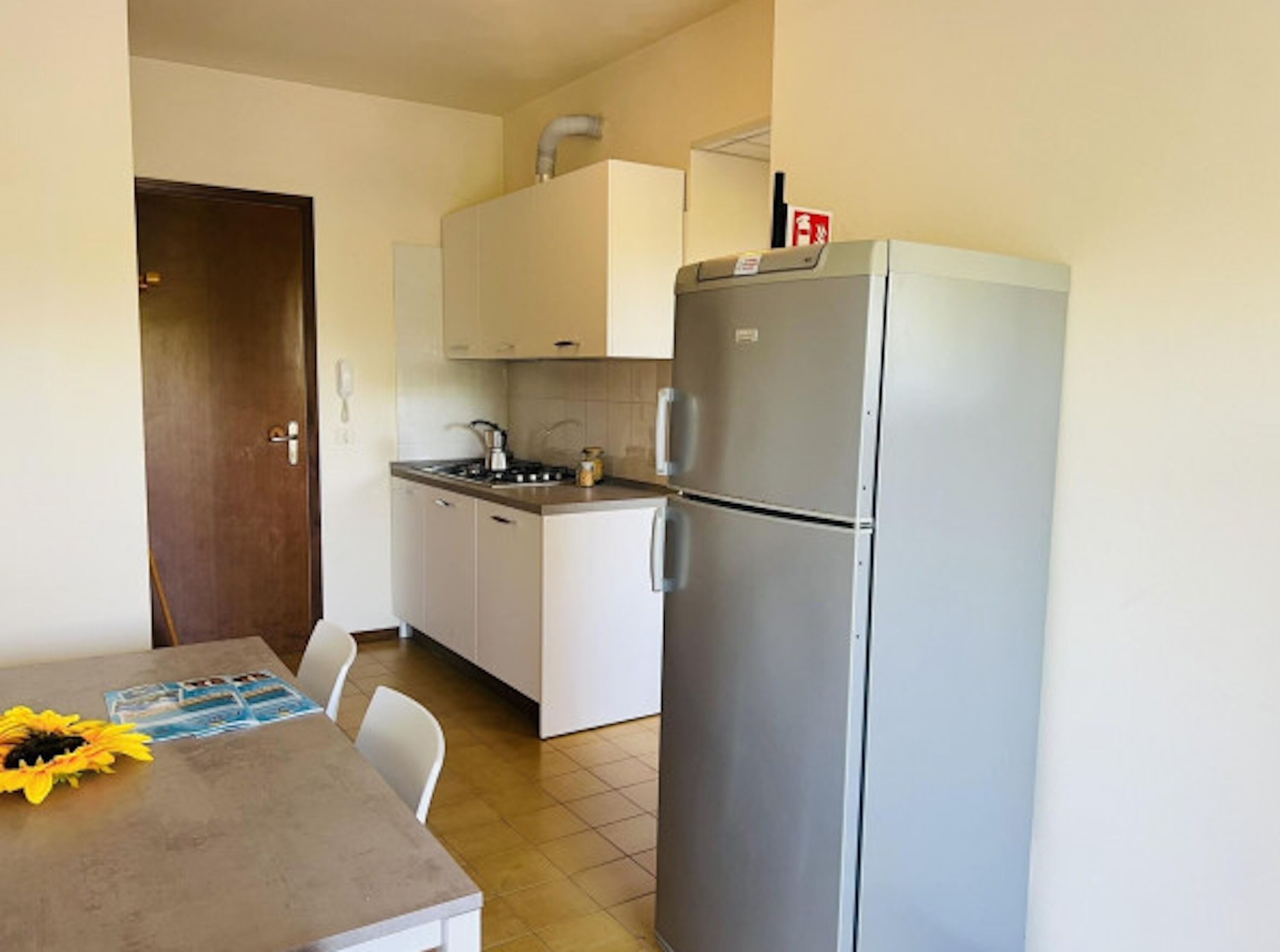 Une cuisine compacte équipée d’un réfrigérateur, d’une cuisinière et d’un évier. On y trouve une porte en bois et une table décorée de motifs de tournesols.
