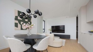 A modern living room with a round dining table, two chairs, a vase with flowers, a large abstract painting, a flat-screen TV, and a sofa.