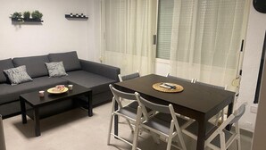 Ein kompakter Wohnbereich mit einer Couch, einem Esstisch und Stühlen. An der Wand befinden sich zwei kleine Regale mit Pflanzen.