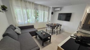 Ein modernes Wohnzimmer mit grauem Sofa, einem Couchtisch und einem Esstisch mit Stühlen. Ein Fernseher ist an der Wand montiert und darüber befindet sich eine Klimaanlage.