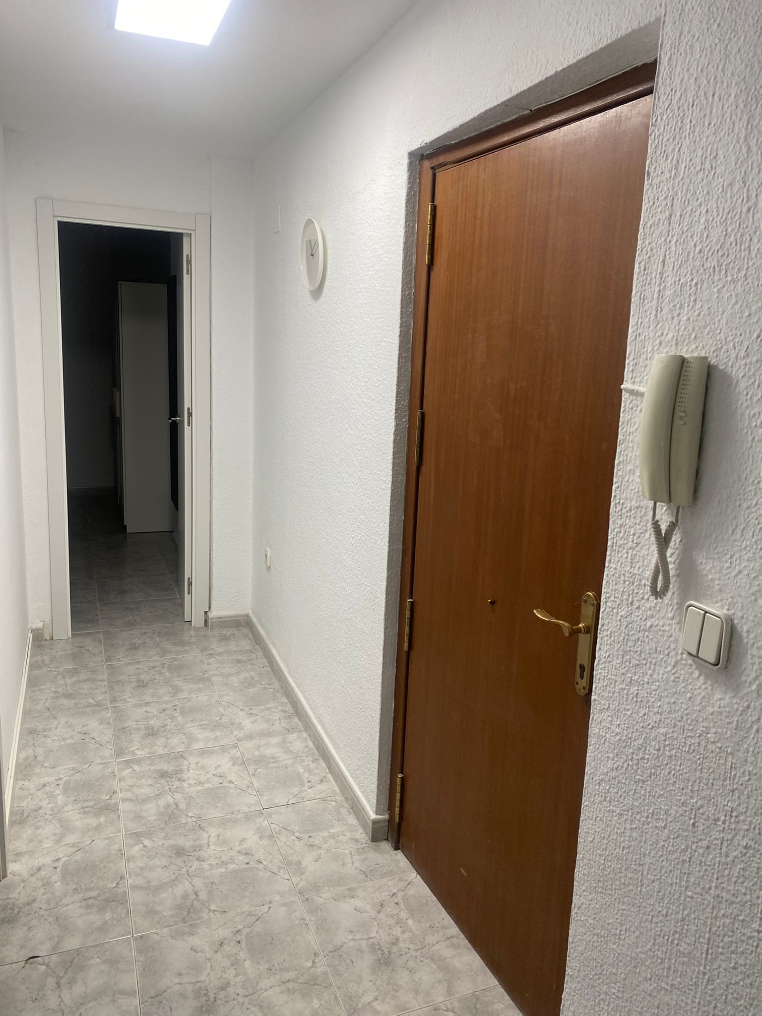 Un pasillo con una puerta de madera, una pared blanca con un termostato y un piso de baldosas.
