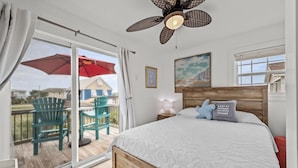 Camera da letto con una testiera in legno, un letto grande con biancheria bianca, un ventilatore a soffitto e vista su un balcone esterno con sedie e un ombrello rosso.