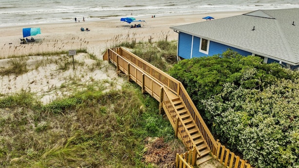 Una casa sulla spiaggia con una scala di legno che conduce a una zona sabbiosa, un edificio blu e l'oceano sullo sfondo.
