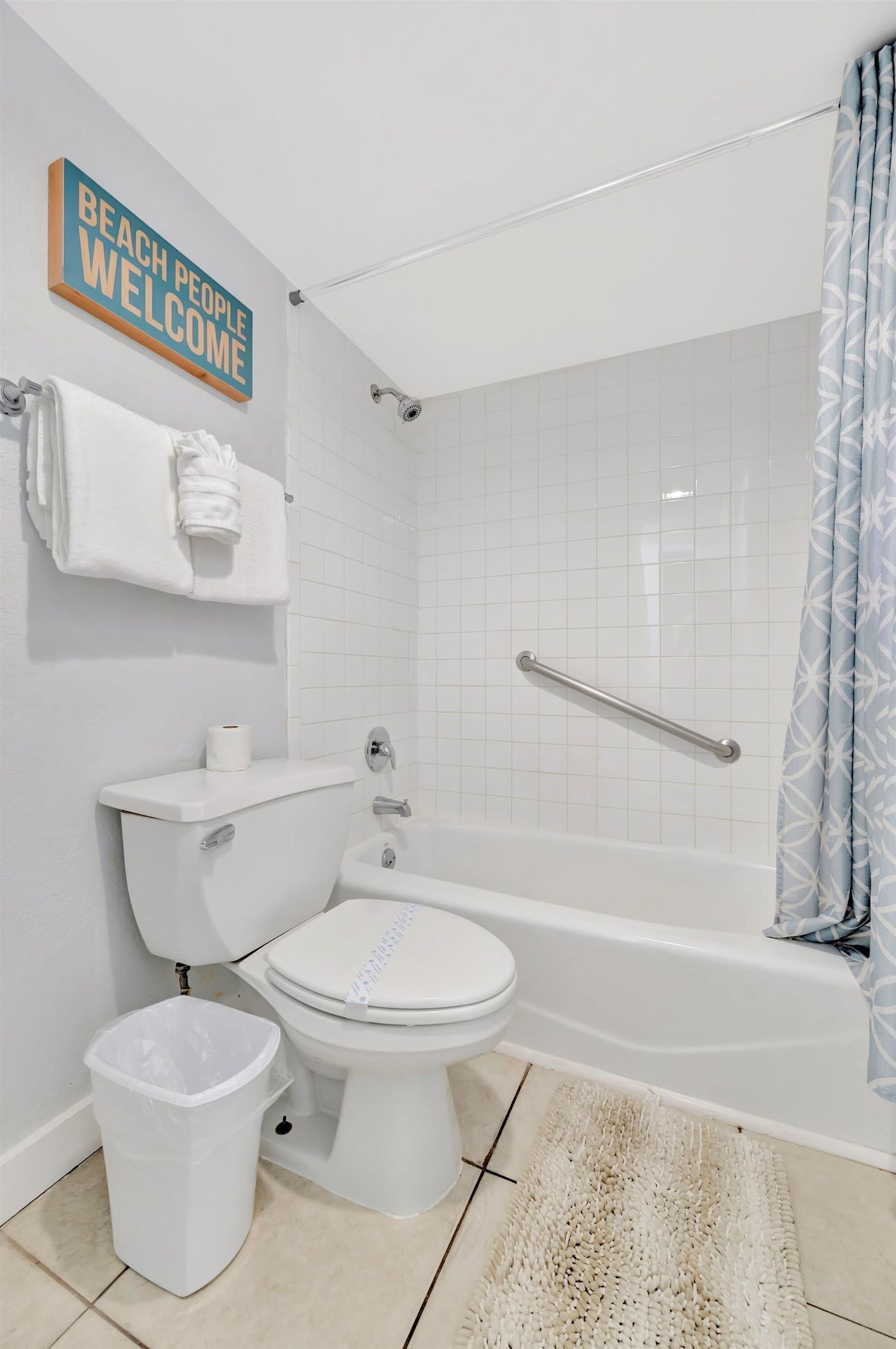 Une salle de bain équipée de toilettes, d’un bidet et d’une baignoire. On y trouve un porte-serviettes avec une serviette et une poubelle. Une pancarte indique : « LES AMANTS DE LA PLAGE SONT LES BIENVENUS ».