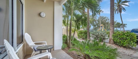 A balcony with white chairs and a small table, overlooking a garden with palm trees and a yellow structure.