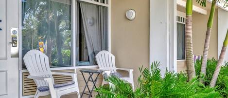 Un balcón con dos sillas blancas, una mesita y vistas a palmeras y un vehículo amarillo a través de la ventana.