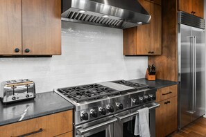 A modern kitchen with stainless steel appliances, wooden cabinets, and a black countertop. - Lavish 6 Bedroom Ski Accessible Home (Big Sky)