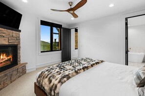 Ein modernes Schlafzimmer mit Kamin aus Stein, einem großen Bett mit Felldecke, Deckenventilator und Blick ins Freie.