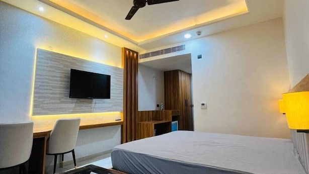 A modern hotel room with a bed, a glass-top table, a desk with a chair, a television mounted on the wall, and a ceiling fan.