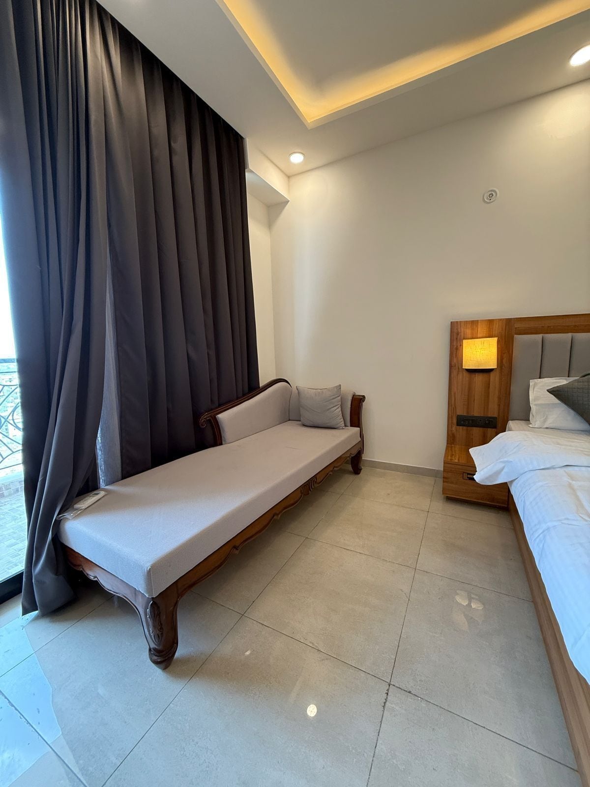 A modern hotel room with a sofa bed, a wooden headboard with a nightstand, and a large window with curtains.