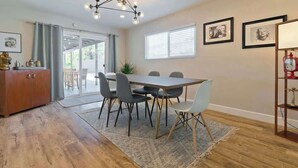 A dining area with a wooden table, grey chairs, and a chandelier. There are framed pictures on the wall and a red clock on a shelf. - Palm Springs Oasis w/ Pool & Spa (Palm Springs)