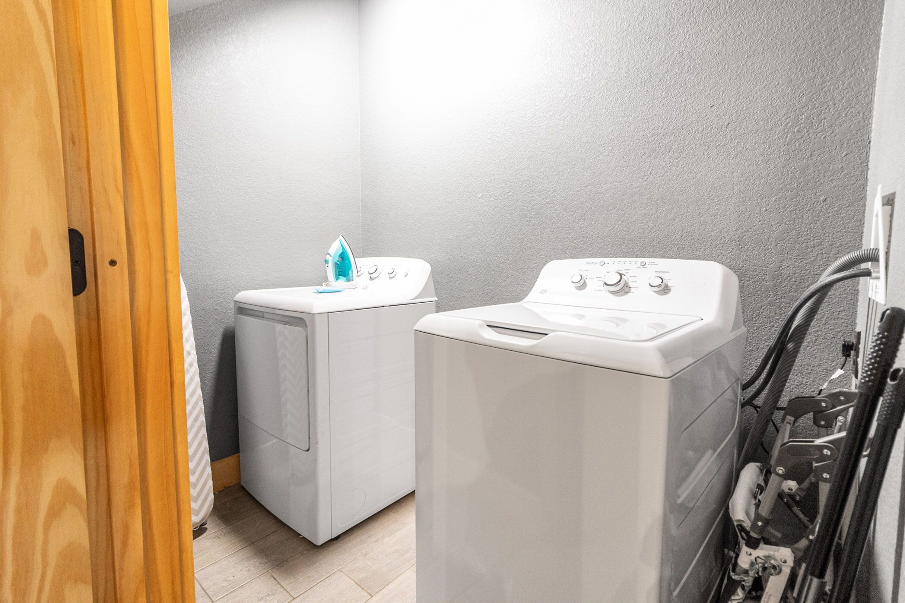 A laundry room with two white washing machines and a dryer, a wooden door, and a tiled floor.