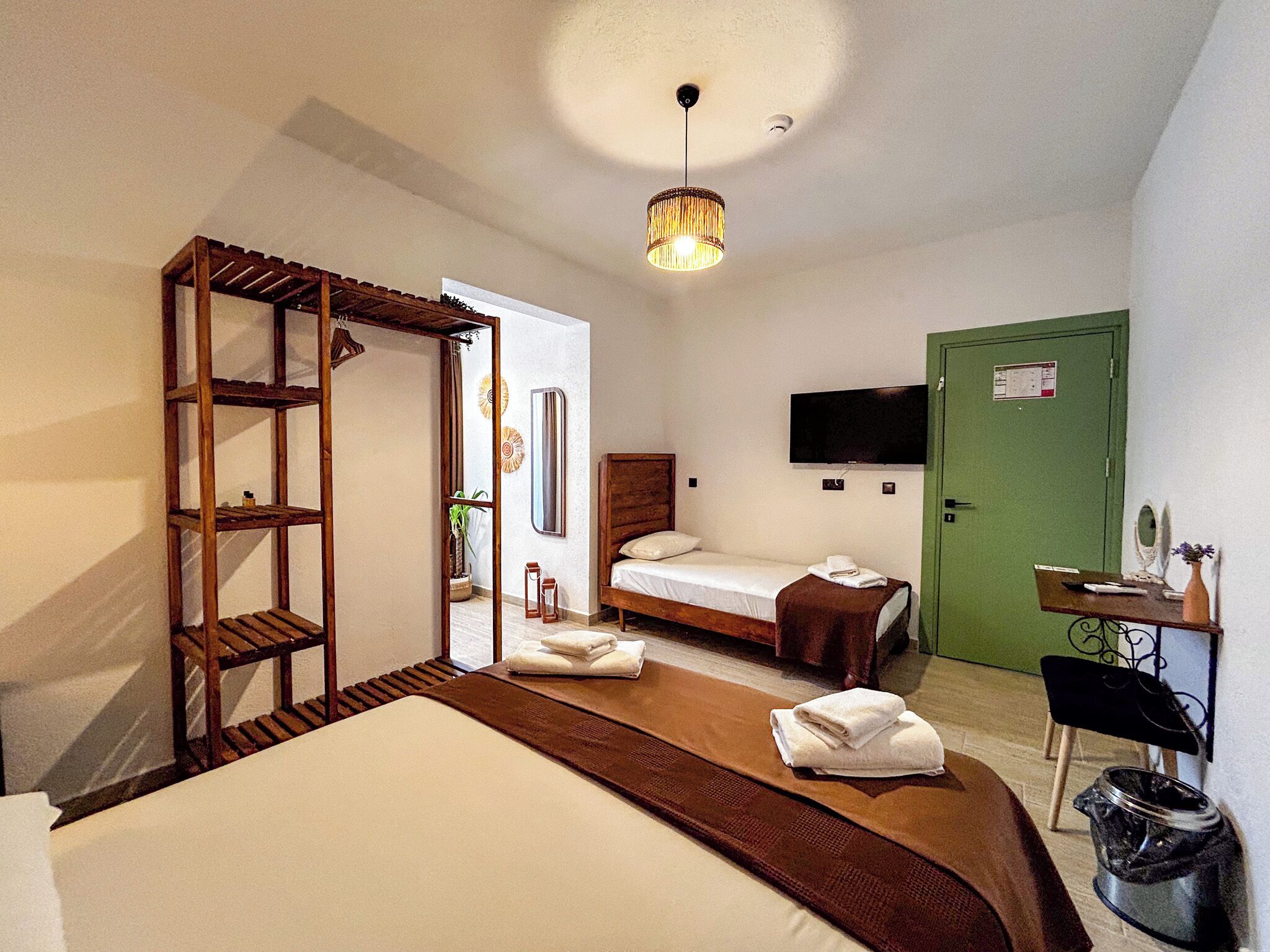A hotel room with a bed, a wooden shelf, a desk with a chair, a television, and a green door.