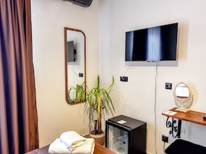 A hotel room with a bed, a TV mounted on the wall, a small refrigerator, a mirror, and a potted plant.