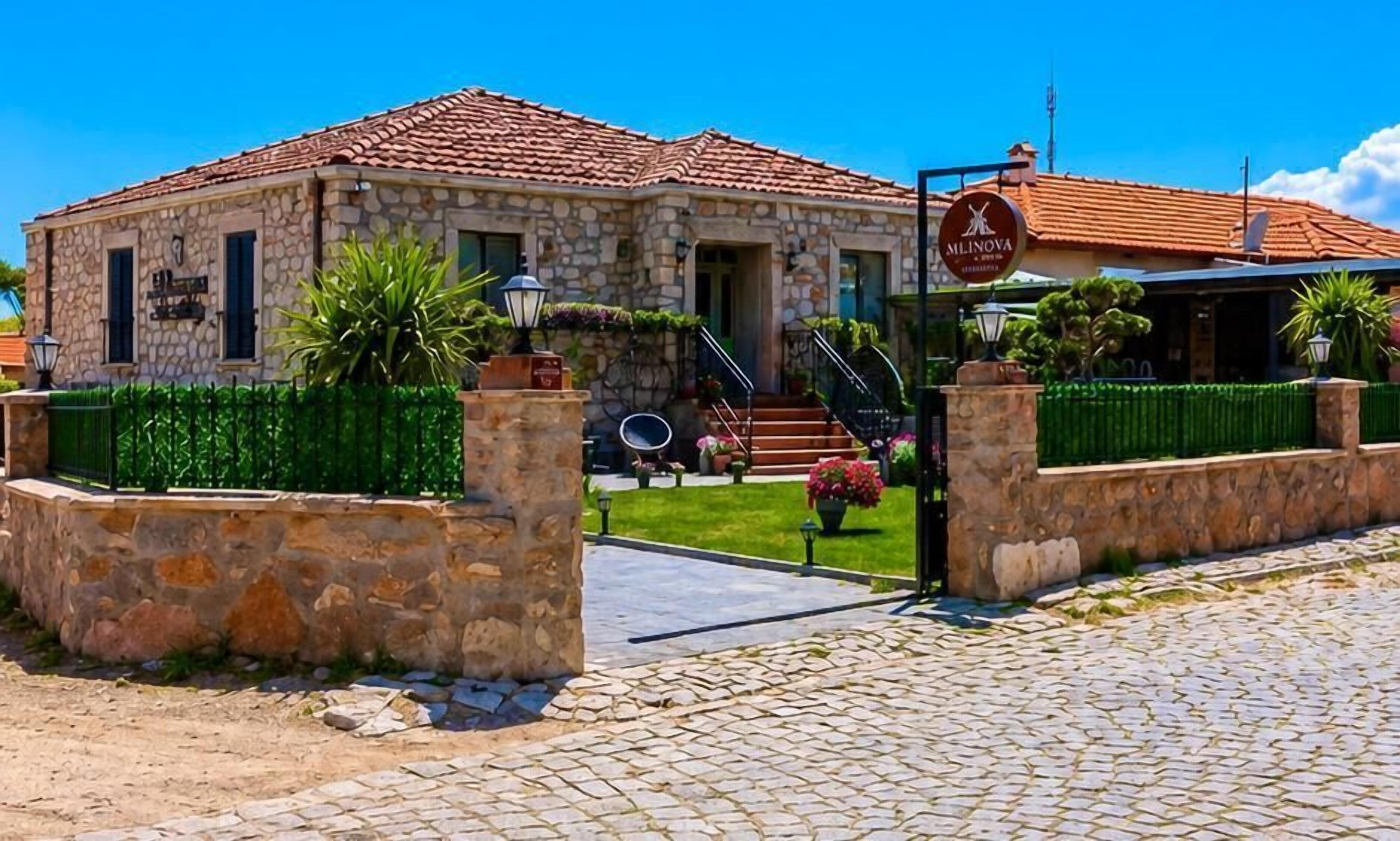 A stone building with a tiled roof, a sign reading 'Mlinova,' and a paved entrance area with a green fence and potted plants.
