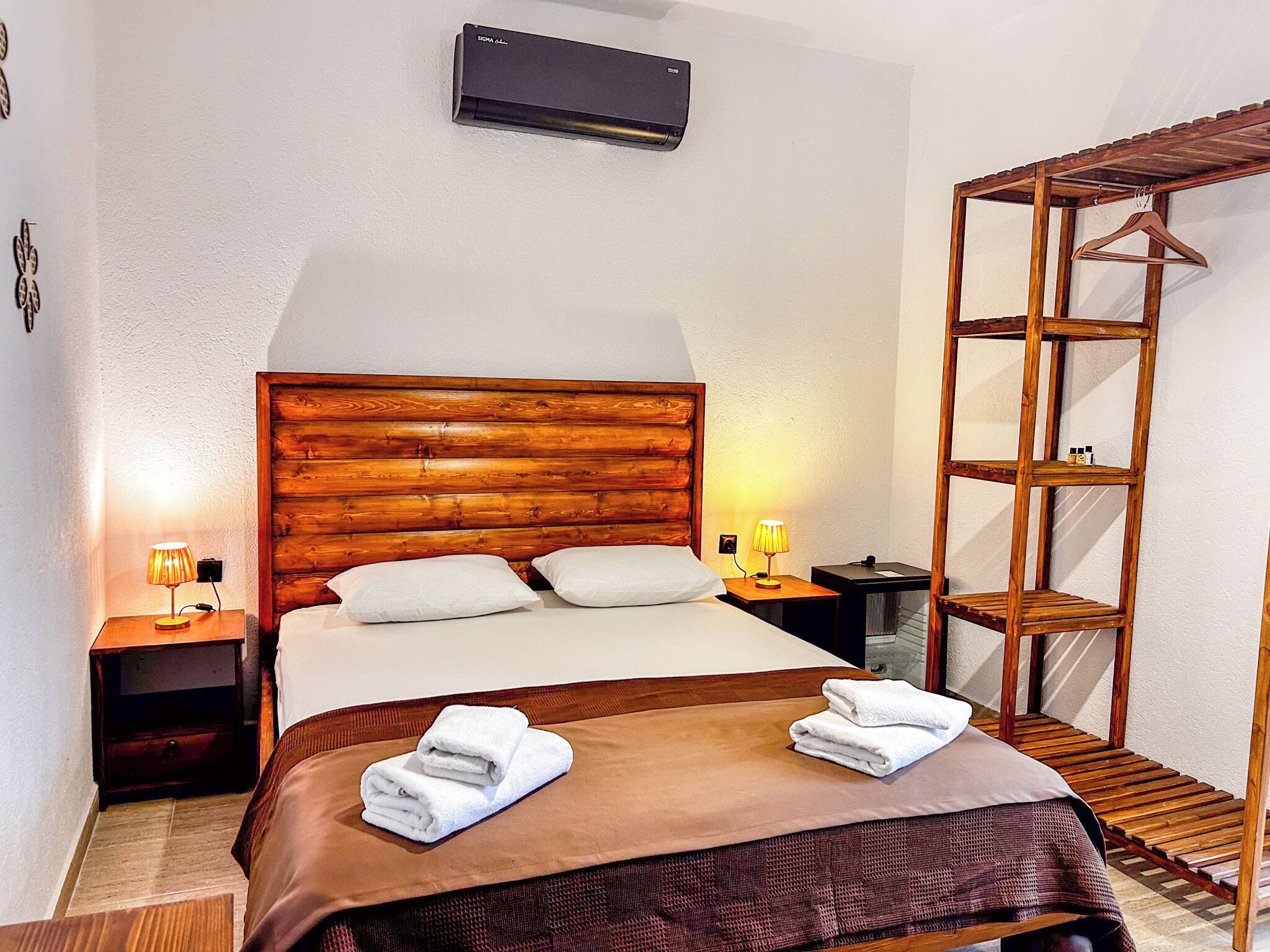 A neatly made bed with a wooden headboard, two bedside lamps, a wooden shelf unit, and a wall-mounted air conditioner.