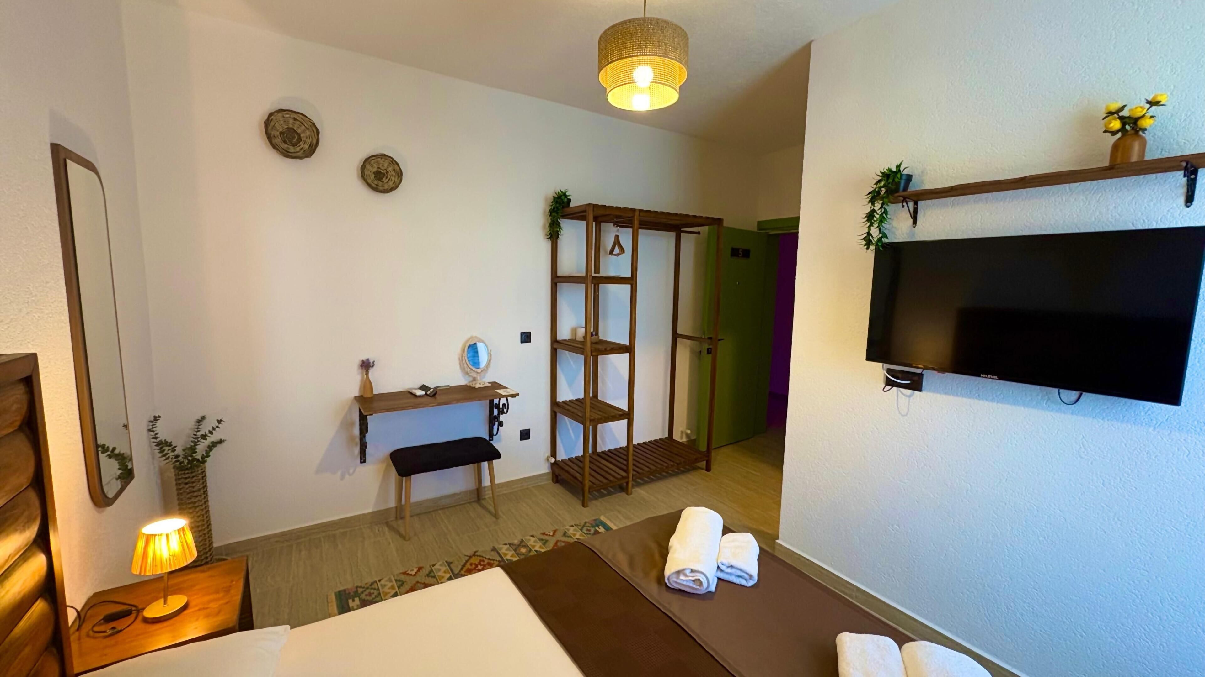A hotel room with a bed, a desk with a mirror, a shelf with decorative items, a television mounted on the wall, and a door leading to another room.