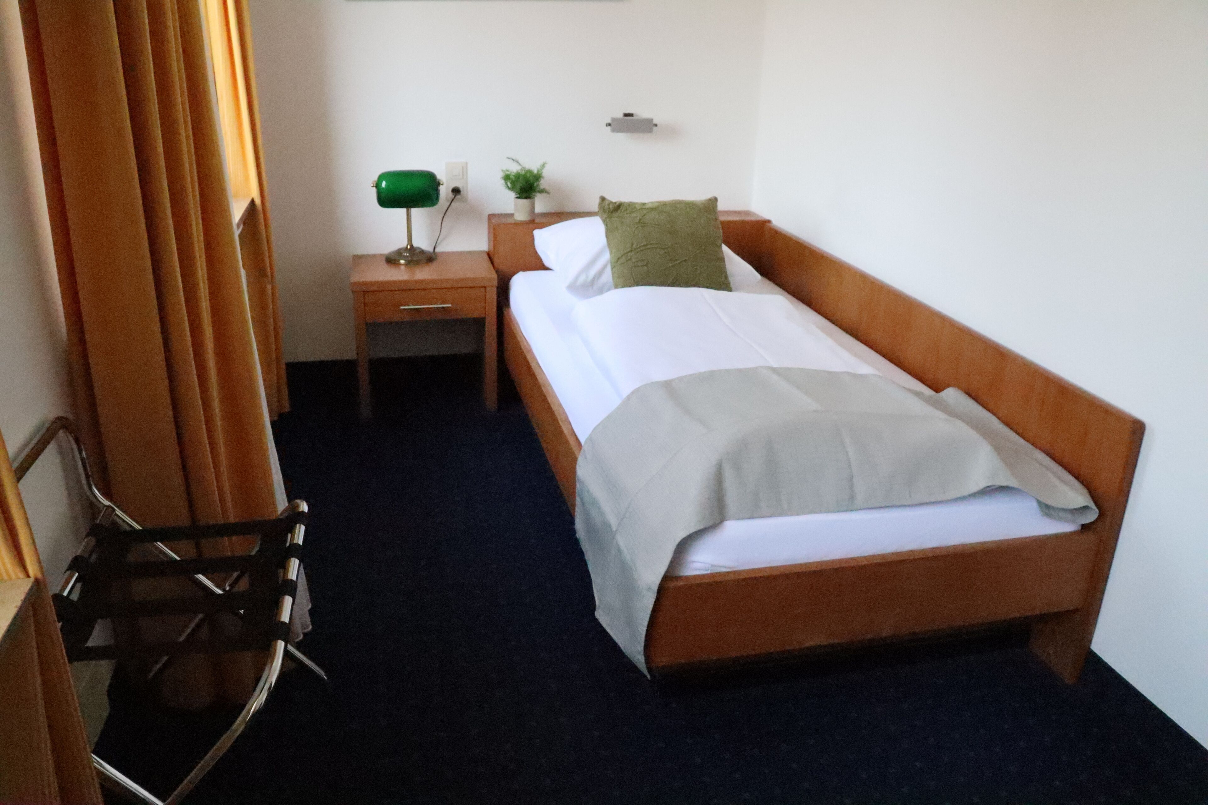 Una camera d'albergo con un letto singolo, un comodino, una lampada, una piccola pianta e una sedia.