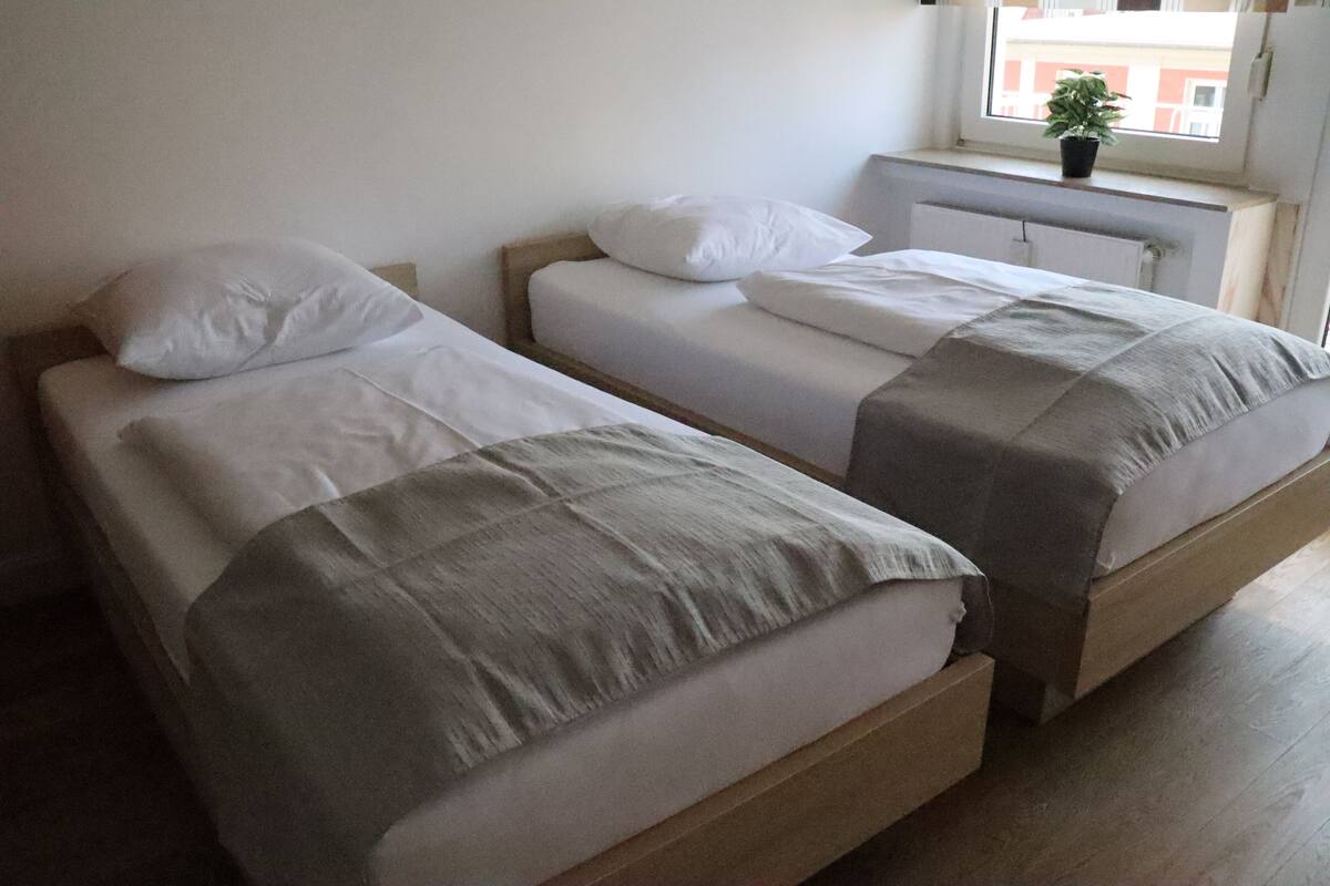 Two single beds with wooden frames and white bedding, a radiator, and a window with a plant.
