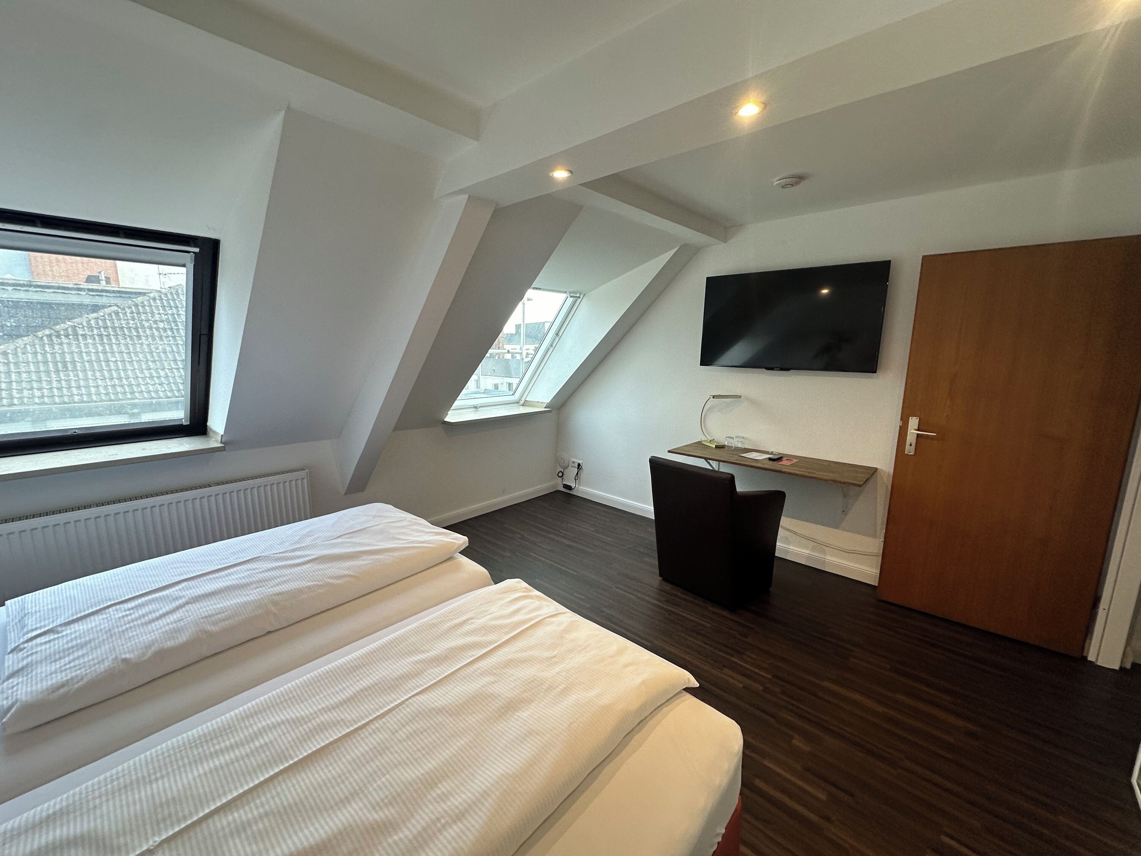 A modern, well-lit room with a bed, a desk, a TV mounted on the wall, and a large window.
