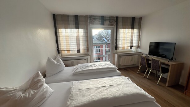 A hotel room with two beds, a desk with a chair, a television, and large windows with curtains. - Hotel Horstmann (Münster)