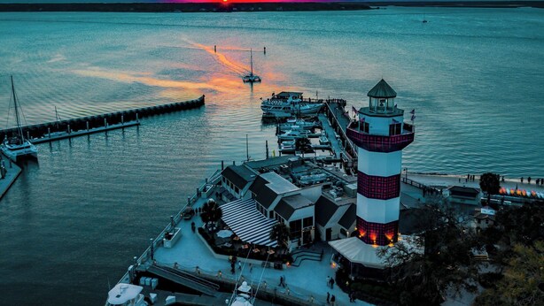 Een vuurtoren met een toren in wit en rood gestreept, een steiger met boten, en een pier die het water in reikt.